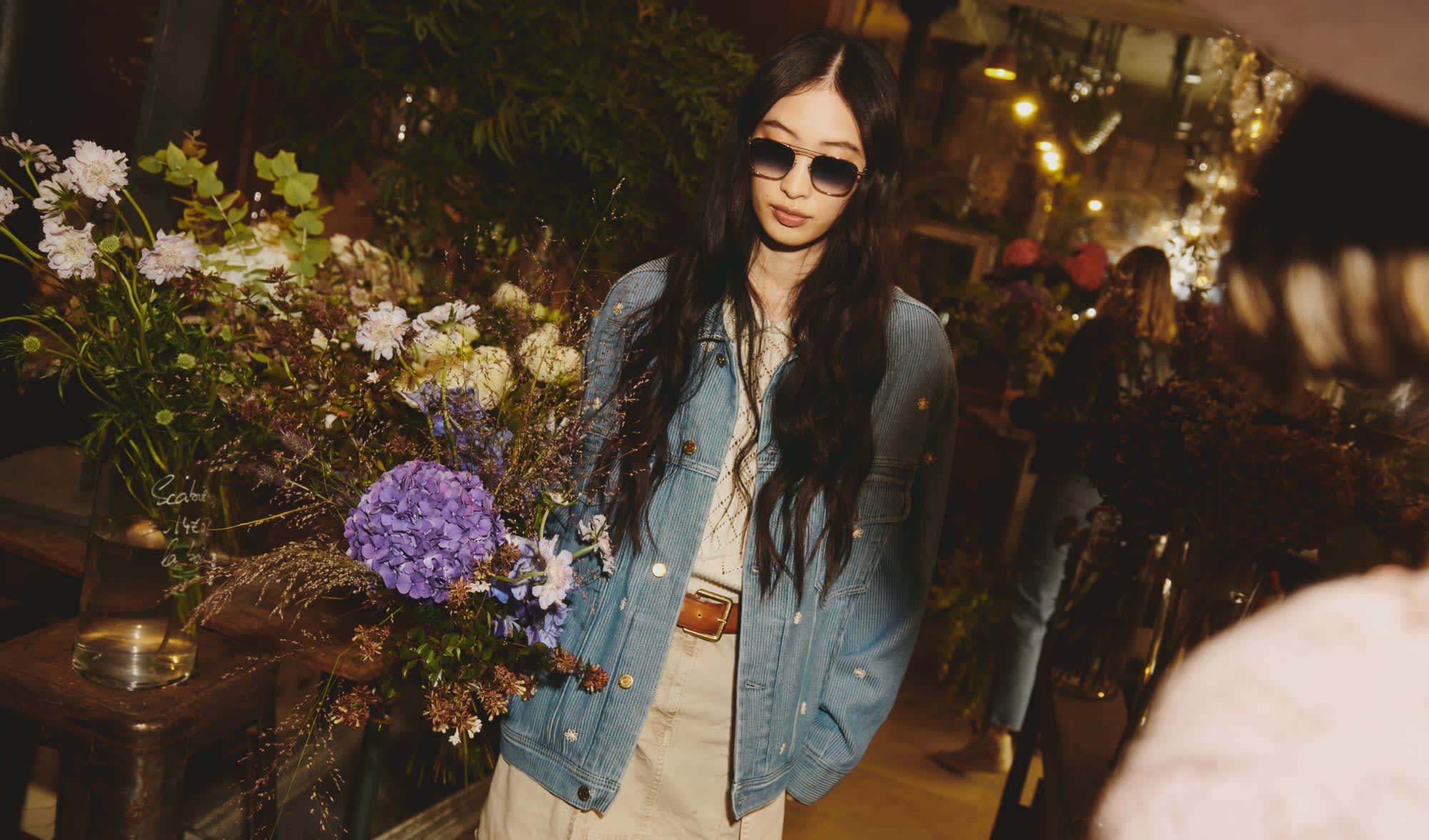woman-with-black-long-hair-in-flower-shop-wearing-denim-jacket-sunglasses-and-beige-skirt